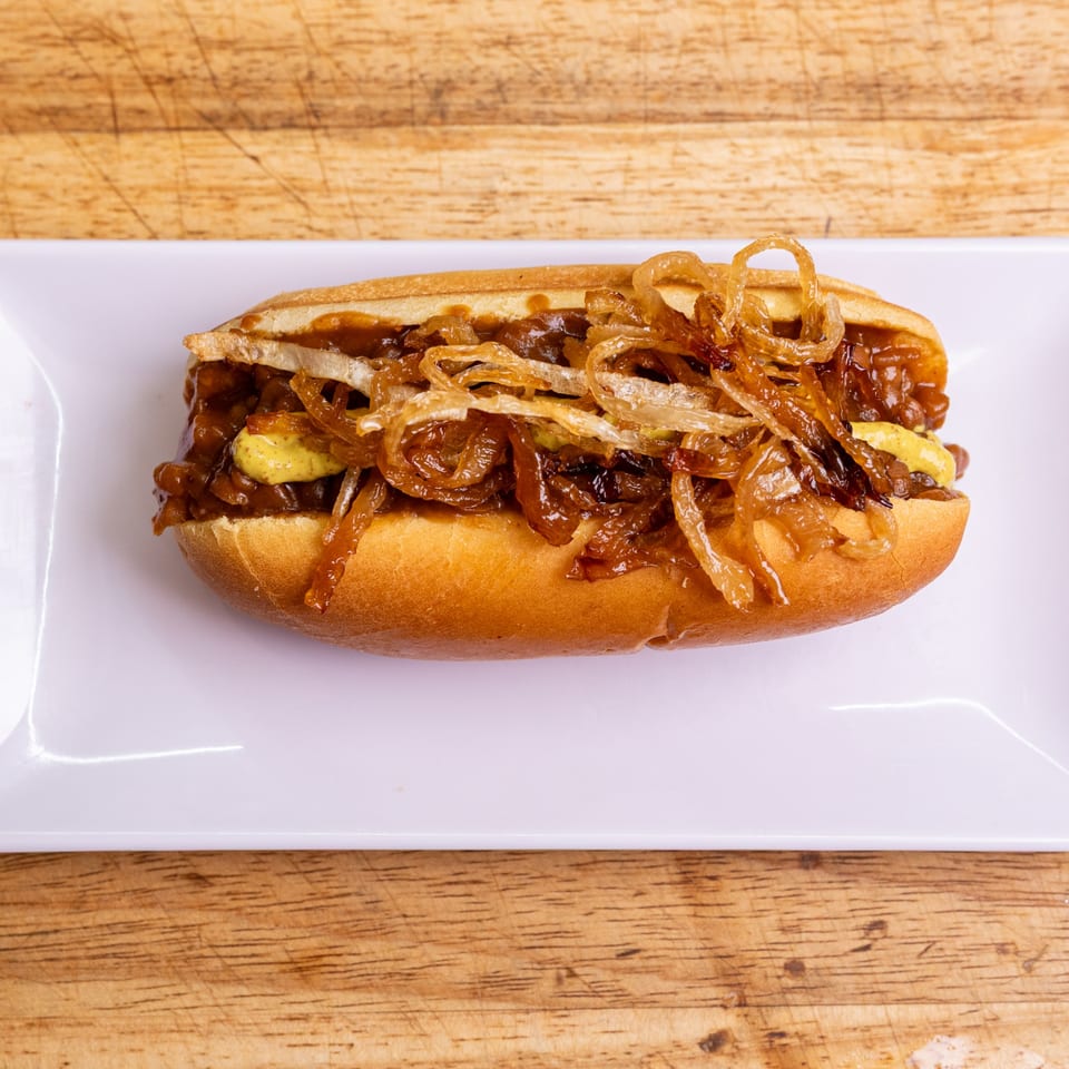 Our Place hot dog with caramelized onions and mustard on a white plate.