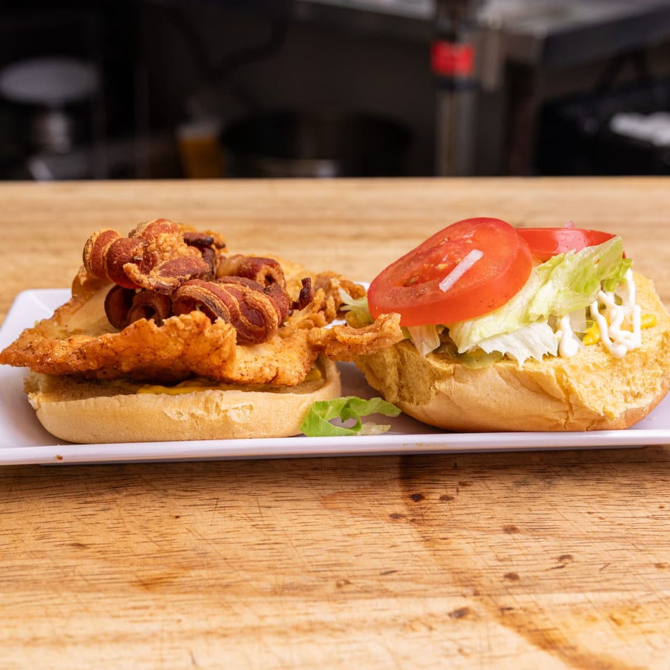 Open-faced fried chicken sandwich with bacon, lettuce, tomato, and mayonnaise on a white plate.