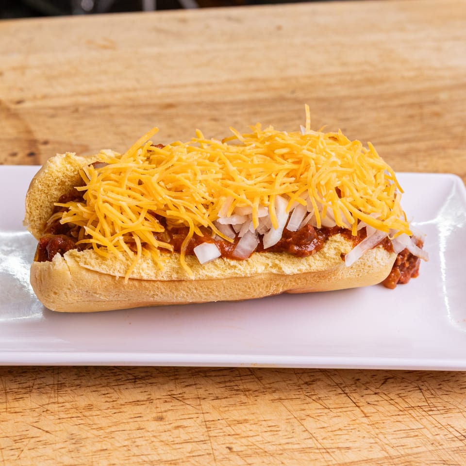 Our Place's chili dog, topped with shredded cheddar cheese and diced white onions, on a white plate.